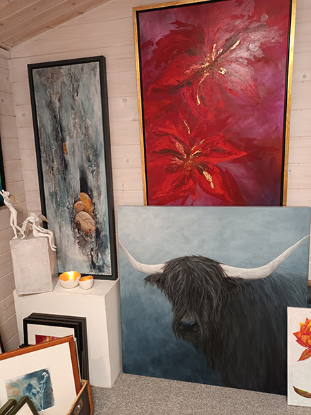 Mehrere Gemälde in einem Raum, darunter ein rotes Blumenbild, ein Porträt eines Hochlandrinds mit langen Hörnern und ein abstraktes Bild mit Steinen, daneben eine Skulptur und kleine Keramikschalen auf weißen Sockeln