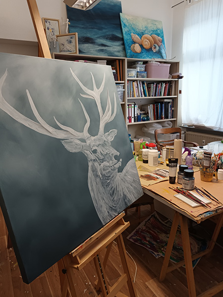 Atelier mit Staffelei, auf der eine Leinwand mit einer halb fertigen Hirschzeichnung steht, im Hintergrund Regale mit Büchern und weiteren Bildern sowie ein Tisch mit Malutensilien