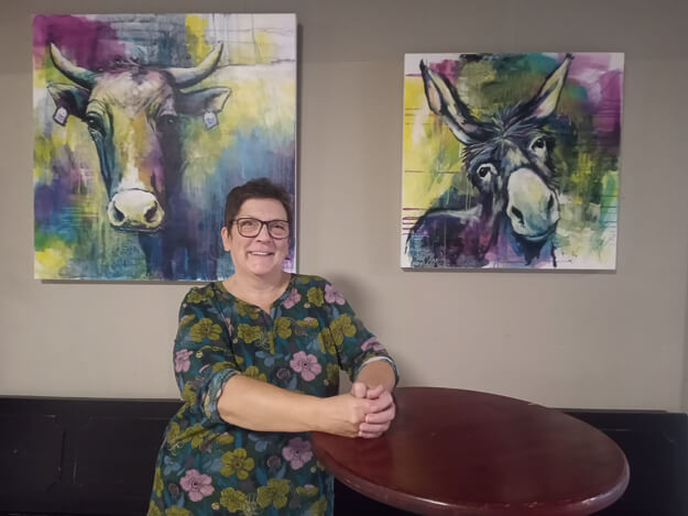 steht vor einer grauen Wand, an der zwei ihrer farbenfrohen Tierporträts hängen: links eine Kuh und rechts ein Esel, beide im expressiven, farbintensiven Stil. Sie trägt ein florales Oberteil und stützt ihre Hände auf einen runden Stehtisch im Vordergrund.