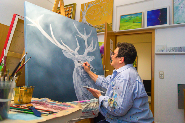 Hildegunde Bode arbeitet in ihrem Atelier konzentriert mit einem Pinsel an einem großformatigen Acrylgemälde eines weißen Hirsches auf dunkelblauem Grund. Im Hintergrund sind weitere farbenfrohe Kunstwerke an den Wänden und auf Staffeleien zu sehen.