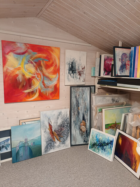 Einblick in eine gemütliche Atelier-Ecke unter einer Holzschräge. An der Wand hängt ein großes, dynamisches abstraktes Gemälde in Rot- und Gelbtönen, daneben ein kleineres schwarz-weißes Werk. Am Boden und auf Regalen sind zahlreiche weitere gerahmte und verpackte Gemälde gelagert, was die kreative Schaffenskraft zeigt.